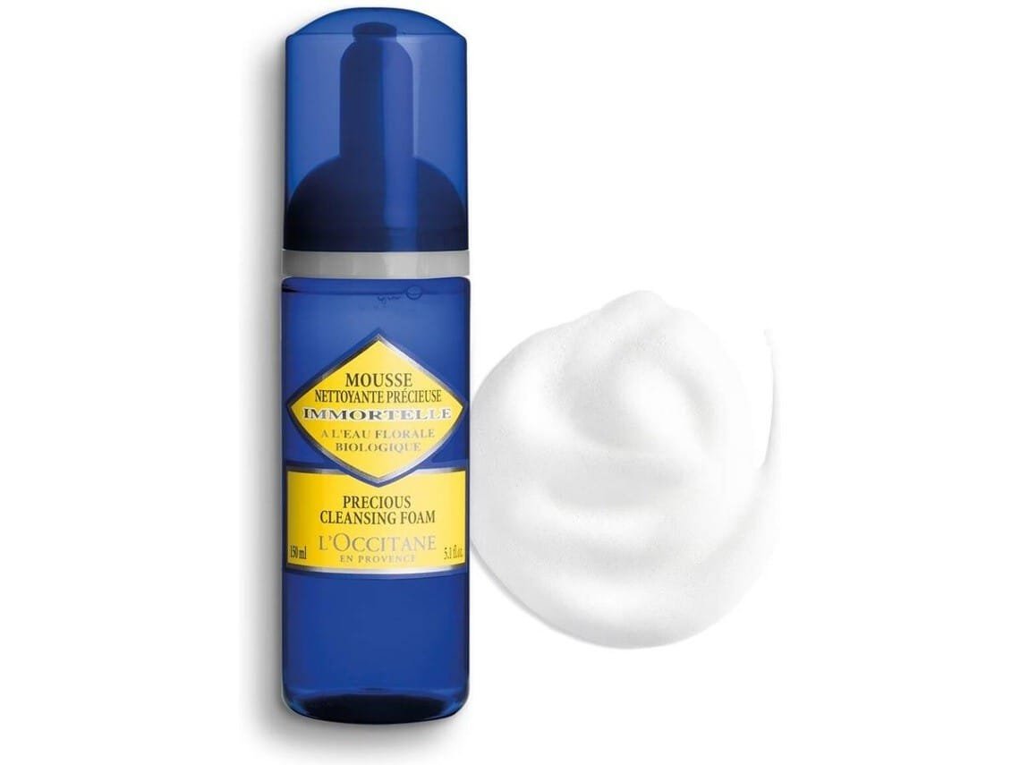 Envase azul con dispensador de la mousse limpiadora L'Occitane Immortelle Precious Cleansing Foam de 150 ml, acompañado de una muestra de espuma blanca, formulada con agua floral de siempreviva orgánica, enriquecida con agua floral orgánica de siempreviva (Immortelle), reconocida por sus propiedades antioxidantes y rejuvenecedoras, esta espuma elimina impurezas, exceso de grasa y restos de maquillaje sin resecar ni agredir la piel. Su textura cremosa y aireada proporciona una experiencia sensorial única que purifica y revitaliza desde el primer uso.