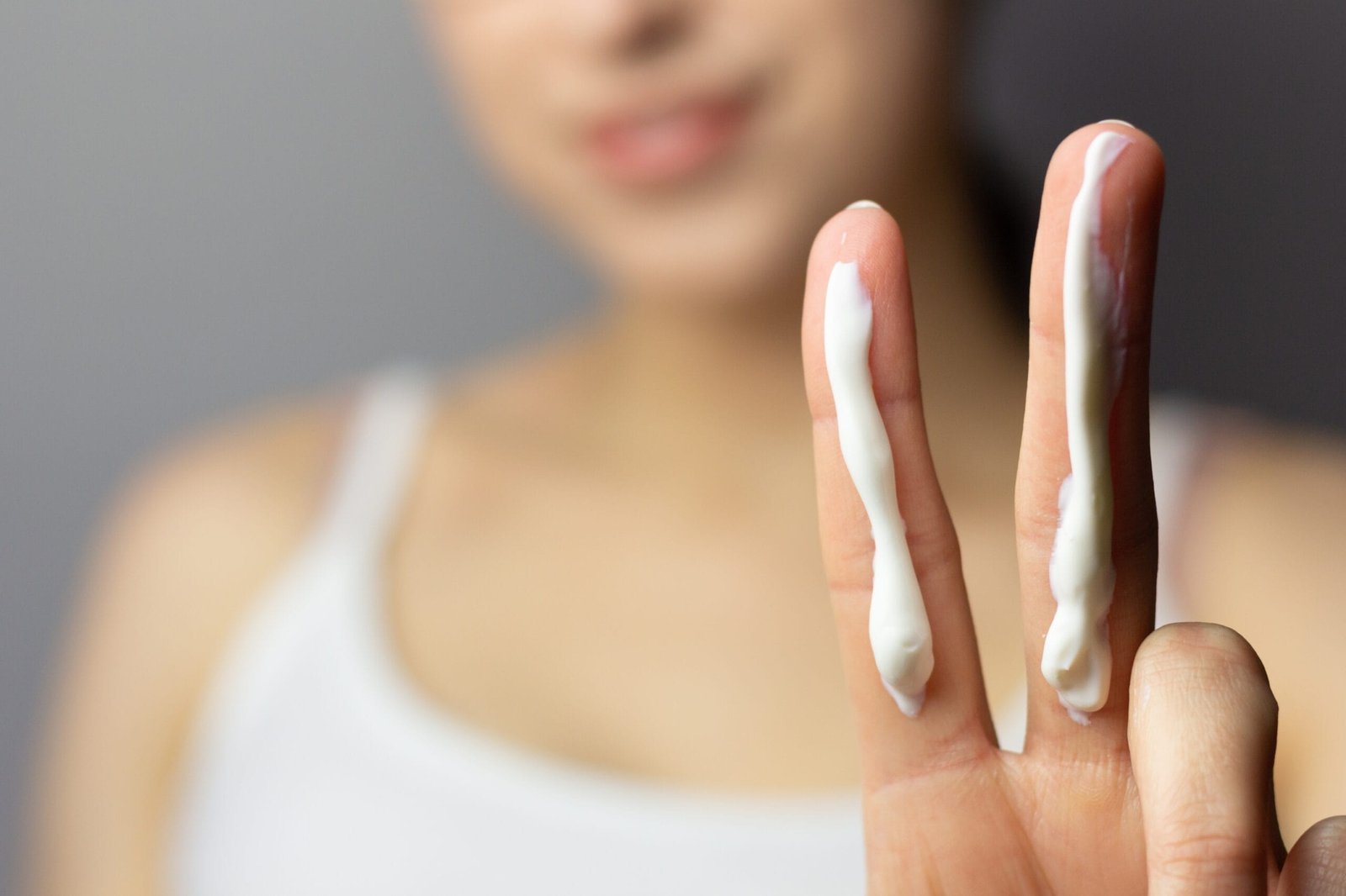 La imagen muestra a una mujer aplicando crema hidratante en sus dedos como parte de su rutina de cuidado de la piel. En primer plano, se destacan dos dedos (índice y medio) con una línea de crema cosmética, ideal para hidratar la piel y mejorar su aspecto. La mujer, que viste una camiseta blanca, aparece con el rostro desenfocado en el fondo, resaltando el enfoque en el tratamiento facial. La escena transmite la importancia de la hidratación facial, el uso de productos de belleza, y el mantenimiento de una piel saludable con cosméticos naturales o de alta calidad. Esta imagen es perfecta para representar temas de belleza, bienestar y cuidado personal, promoviendo el uso de cremas nutritivas y tratamientos para la piel.
