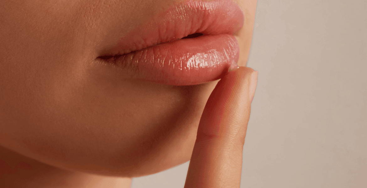 Boca femenina con los labios ligeramente entreabiertos y maquillados con brillo labial. Un dedo índice toca suavemente el labio inferior aplicando crema labial. La piel luce suave y bien cuidada, y el fondo neutro rosado resalta los detalles del rostro.
