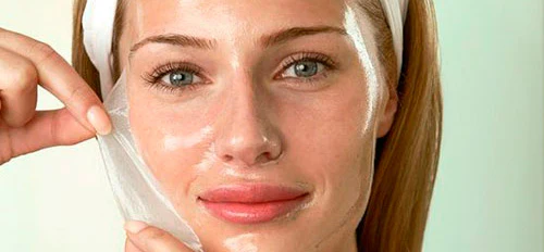 Mujer joven retirando una mascarilla facial transparente de su rostro, con una banda blanca en el cabello.