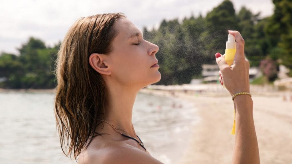 Mujer con el cabello mojado rocía un producto en spray sobre su rostro mientras se encuentra en una playa. Con los ojos cerrados y expresión relajada, disfruta del rocío refrescante bajo la luz natural del día. El fondo muestra un paisaje costero con arena clara, agua tranquila y vegetación frondosa. La escena transmite una sensación de bienestar, autocuidado y conexión con la naturaleza, sugiriendo el uso de un producto como protector solar o agua termal.