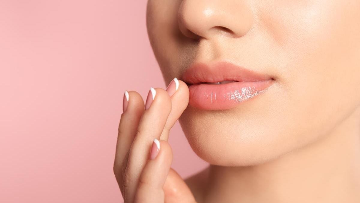 La imagen muestra unos labios hidratados, con un sutil brillo natural, resaltando su suavidad y apariencia saludable. La piel luce radiante y bien cuidada, con un fondo rosado que aporta un toque de feminidad y elegancia. La persona está aplicando un producto, lo que sugiere una rutina de cuidado labial. La estética general transmite una sensación de belleza natural, hidratación y bienestar.