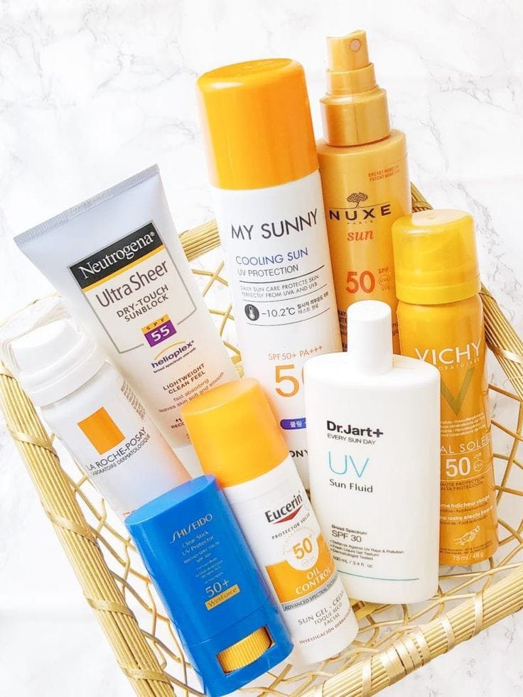 Cesta de mimbre con una selección variada de protectores solares de diferentes marcas reconocidas, como Neutrogena, My Sunny, Nuxe, Vichy, La Roche-Posay, Shiseido, Eucerin y Dr. Jart+, todos con factor de protección solar SPF 50 o superior, en diferentes formatos como spray, fluido, barra y loción.