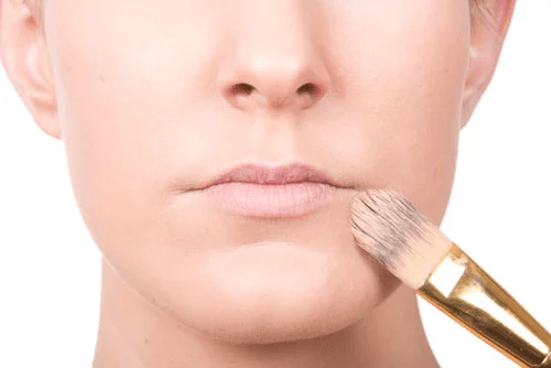 Aplicación de base de maquillaje en labios con brocha, preparación para maquillaje profesional en rostro claro.