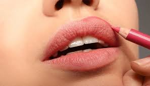 Mujer delineando sus labios con un lápiz labial rosa, mientras mantiene la boca entreabierta, se ve una piel perfecta y suave.