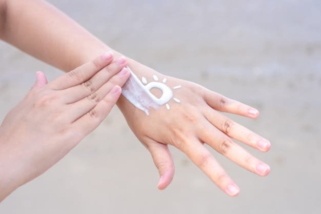 Una mano extiende protector solar sobre el dorso de la otra, donde previamente se ha dibujado con la crema un pequeño sol con rayos alrededor, simbolizando protección solar. La imagen está tomada en un entorno exterior con fondo desenfocado que sugiere una playa o zona costera. La escena transmite el concepto de cuidado personal, prevención del daño solar y hábitos saludables relacionados con la exposición al sol.