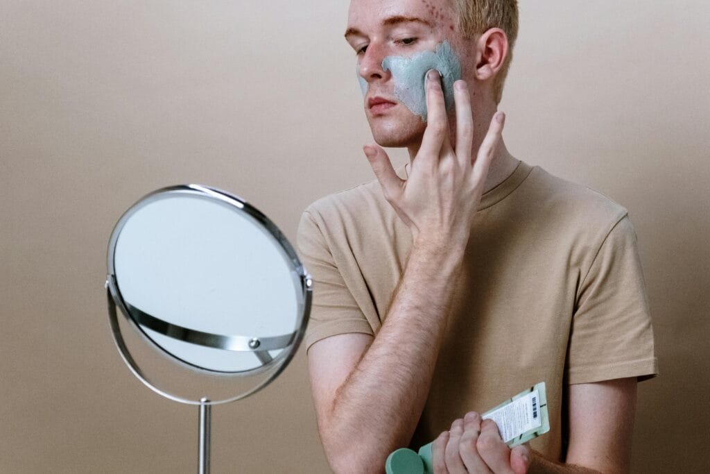 La imagen muestra a un hombre aplicando una mascarilla facial de color verde en su rostro, como parte de su rutina de skincare para tratar acné, piel grasa y marcas en la piel. Sostiene un tubo de crema facial mientras se observa en un espejo de aumento, reflejando el proceso de cuidado de la piel y el uso de productos cosméticos específicos. Lleva puesta una camiseta beige, en un fondo neutro, lo que enfatiza la escena de autocuidado y bienestar.