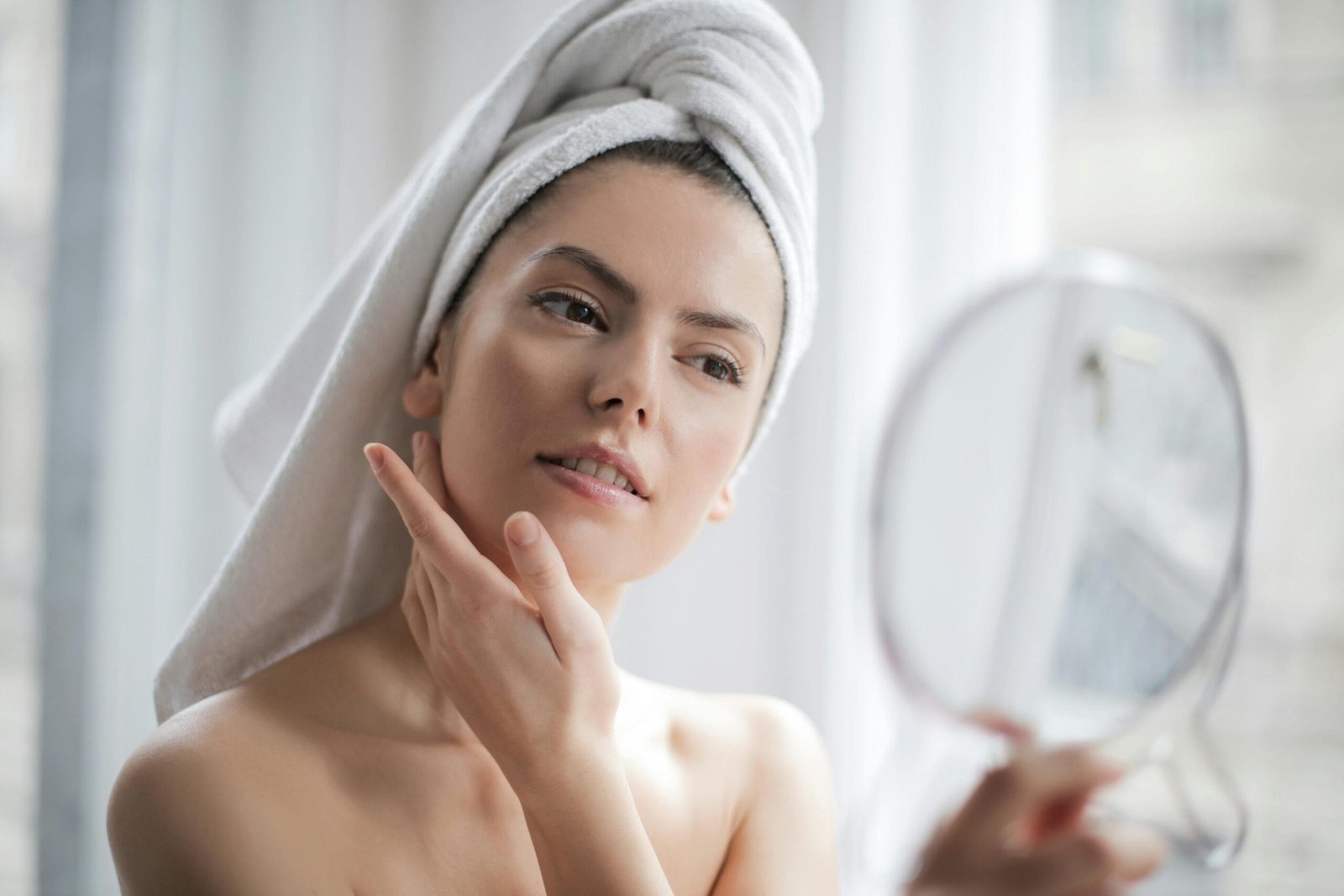 Mujer con piel radiante y natural, con una toalla blanca sobre la cabeza, reflejando una rutina de skincare. Su expresión es relajada y confiada mientras se mira en un espejo de mano, destacando la importancia del autocuidado y la belleza saludable. La luz natural resalta su rostro hidratado, evocando frescura y bienestar. La escena transmite un mensaje de cuidado facial, rutina diaria, y autoestima, con un ambiente de tranquilidad y spa en casa.