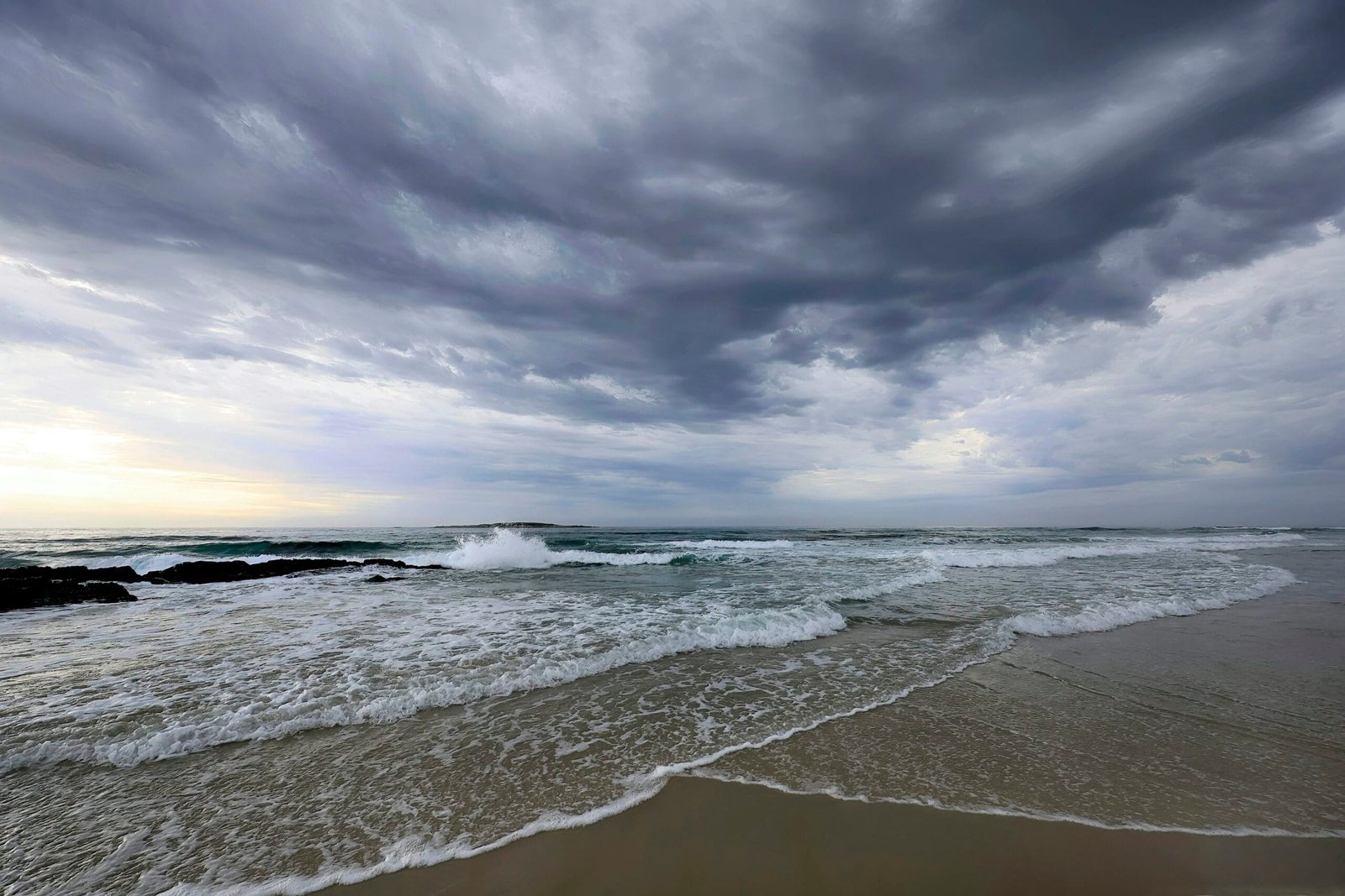 Playa con olas rompiendo suavemente sobre la orilla bajo un cielo cubierto de nubes densas y grises. La escena transmite una atmósfera dramática y serena a la vez, con la luz del amanecer o atardecer asomando sutilmente en el horizonte, iluminando tenuemente el mar agitado y la arena húmeda. La combinación de elementos naturales crea un contraste entre la fuerza del océano y la calma previa a una posible tormenta.