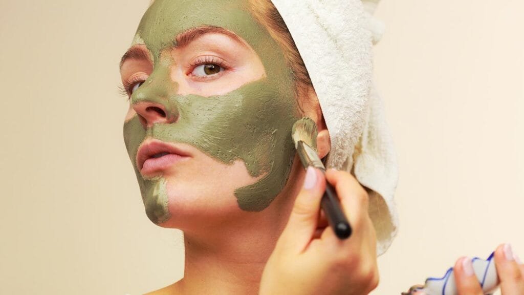 Mujer aplicando mascarilla facial verde con brocha sobre su rostro. Lleva el cabello envuelto en una toalla blanca, mostrando una rutina de cuidado de la piel. Ideal para piel grasa o mixta. Fondo neutro que resalta el tratamiento facial. Imagen perfecta para artículos de belleza, mascarillas naturales, rutinas de skincare y tips para el cuidado del rostro como desinflamar rapido un grano
