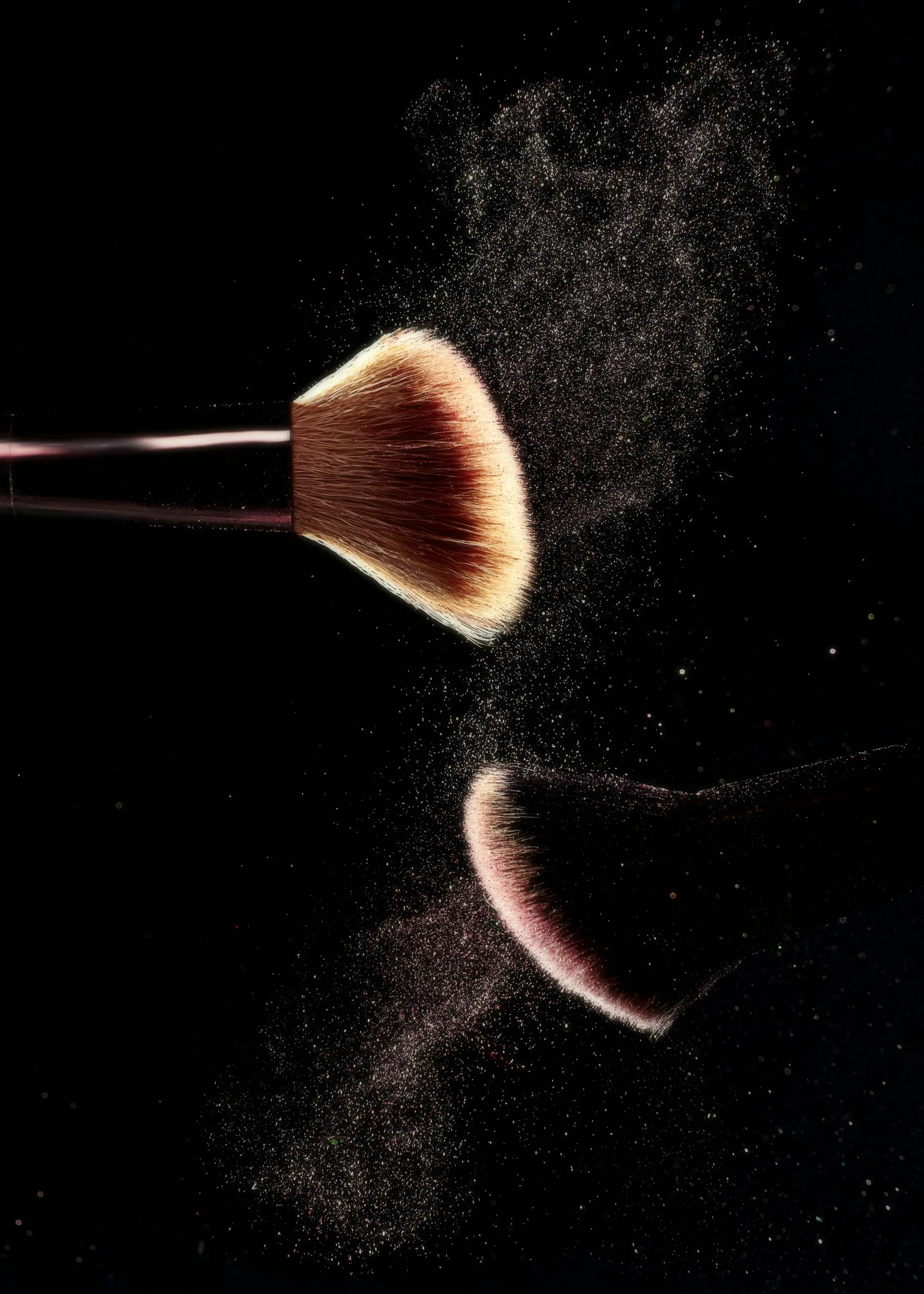 Imagen artística de una brocha de maquillaje profesional soltando partículas de polvo cosmético sobre un fondo negro. Suavidad de las cerdas y la calidad del pincel, ideal para aplicar polvos compactos, iluminadores o rubor. El contraste entre el fondo oscuro y el brillo del polvo flotando en el aire transmite elegancia, detalle y precisión. Consejos y trucos de maquillaje para maquillar tu rostro según su forma,