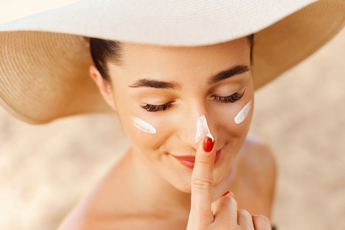 Mujer joven aplicando protector solar en su rostro mientras disfruta de un día soleado en la playa. Lleva un sombrero de ala ancha para protegerse del sol, tiene la piel bien cuidada y marcas visibles de crema solar en la nariz y mejillas. Imagen ideal para artículos sobre protección solar facial, cuidado de la piel en verano y prevención del daño solar.