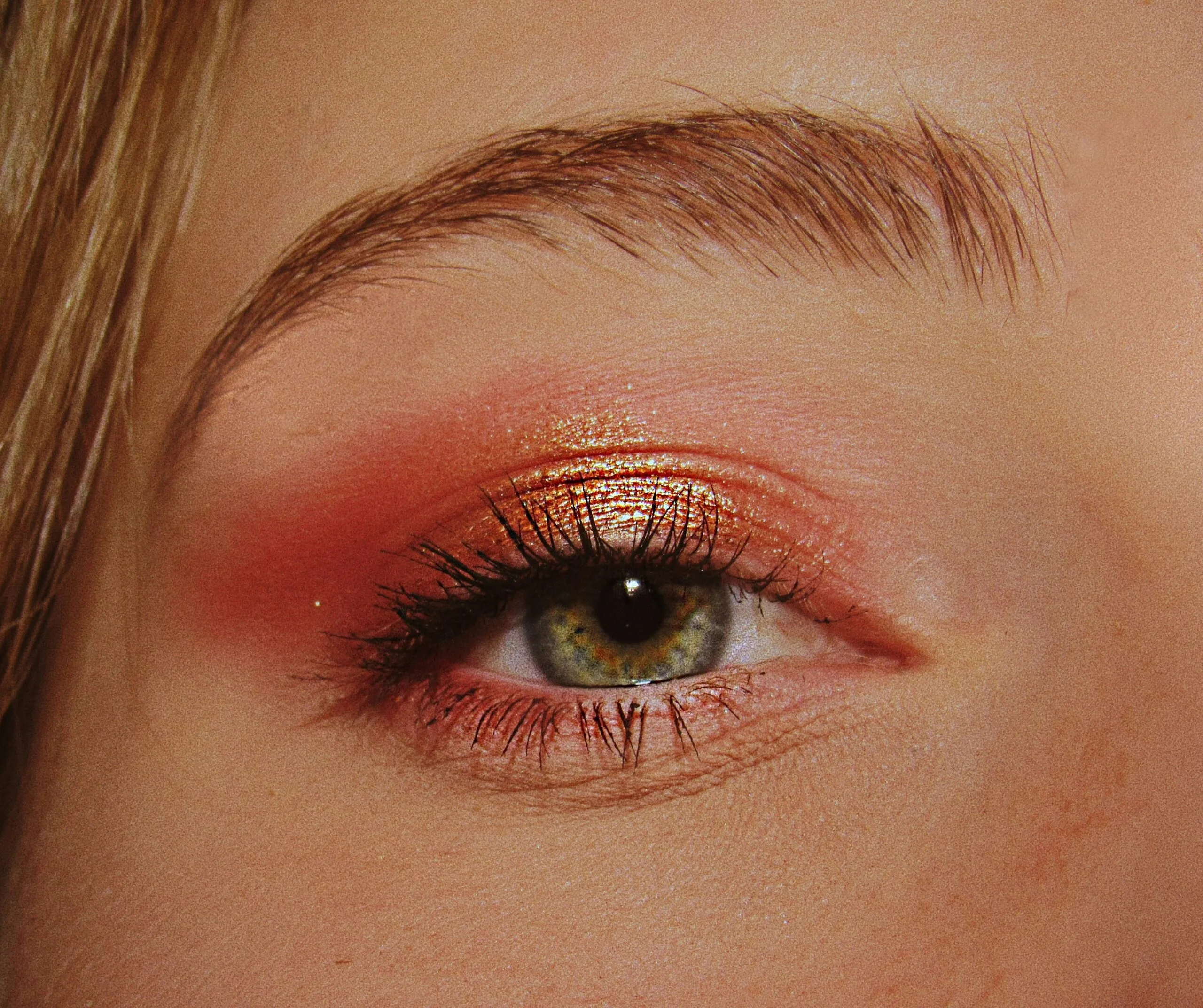 Ojo femenino maquillado con sombra de ojos en tonos cálidos, incluyendo dorado metálico y naranja coral, ideal para resaltar ojos verdes. El maquillaje profesional resalta la mirada con un difuminado perfecto y acabado brillante. Las pestañas están definidas con máscara negra, aportando volumen y longitud. La ceja bien delineada enmarca el ojo, completando un look moderno y sofisticado.